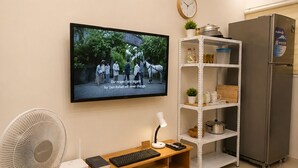 A modern living room with a flat-screen TV mounted on the wall, a wooden entertainment unit with a keyboard and mouse, a standing fan, and a white shelving unit with kitchen items. - Tsinoy Villa (Pandi)