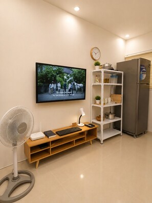 Un salon moderne avec un téléviseur à écran plat fixé au mur, un meuble de rangement en bois comprenant un clavier et une souris, un ventilateur sur pied et une étagère blanche contenant des ustensiles de cuisine.