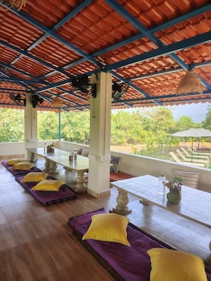 A covered outdoor seating area with wooden tables and benches, yellow cushions, and a view of greenery. - 360 Agonda (Canacona)