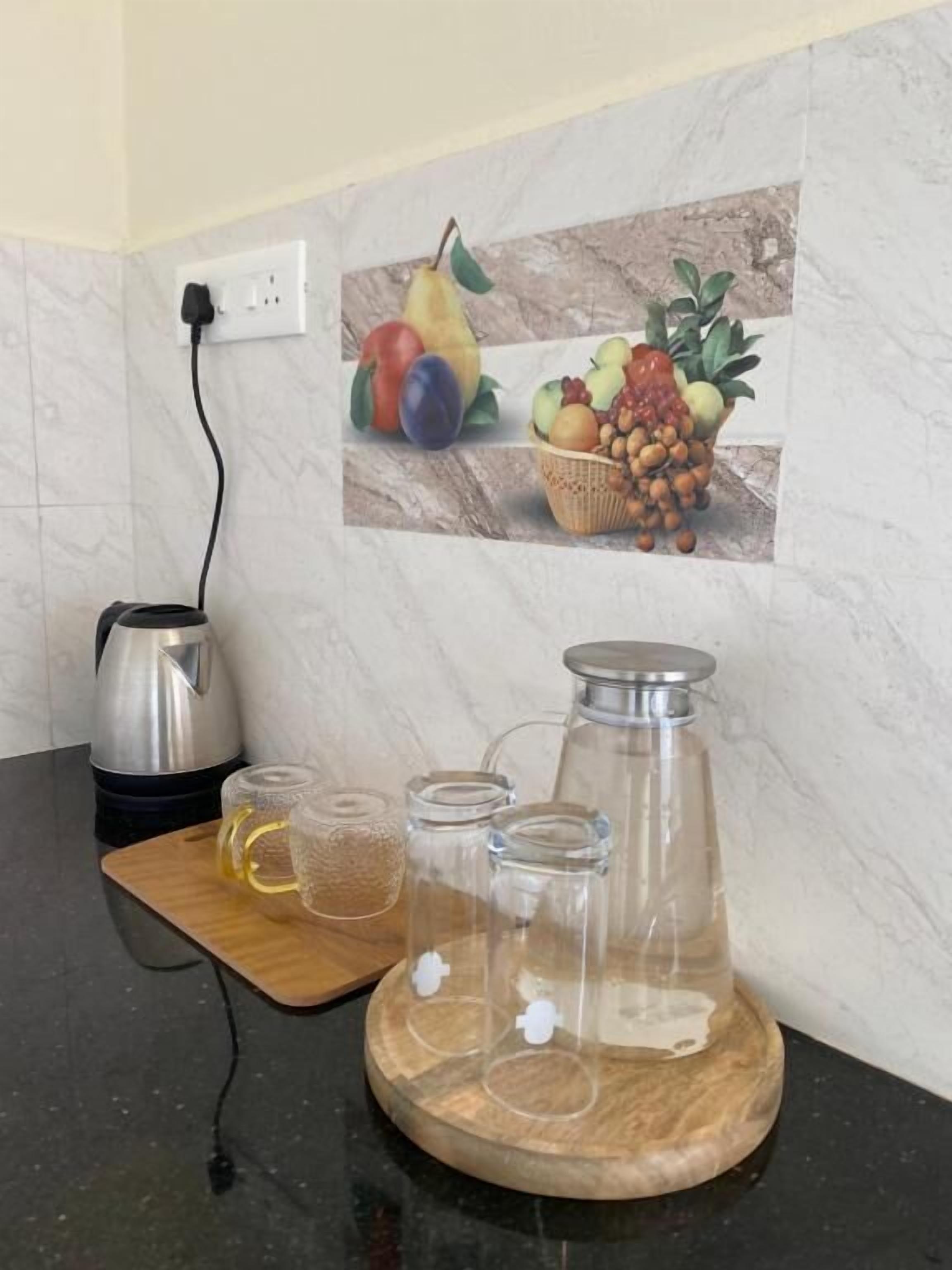 Una cuina amb un taulell, una cafetera, un bullidor i un decantador de vidre. Hi ha una taula de tall de fusta amb un pot de mel i un envasament de vidre. Al mur, hi ha un quadre de fruita i verdura.