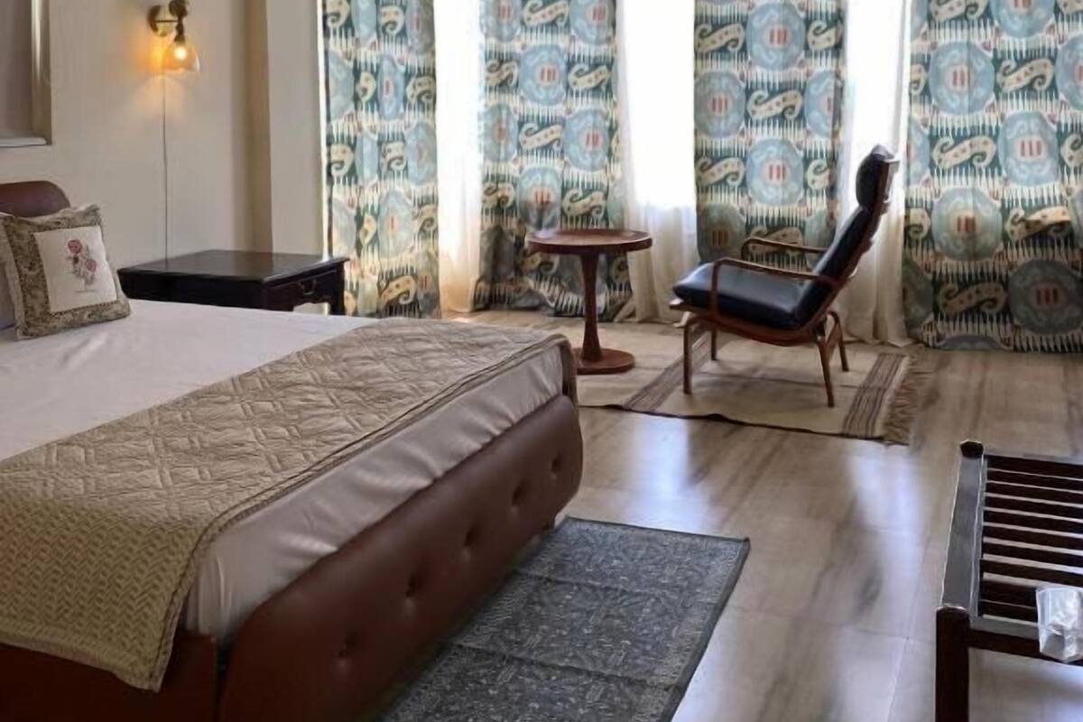 A bedroom with a bed, a chair, a small table, a ceiling fan, and patterned curtains.