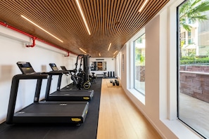 A gym with treadmills, exercise equipment, and a view of a brick wall and greenery outside. - Ocean's Edge Suite at 42 Eilat (Eilat)