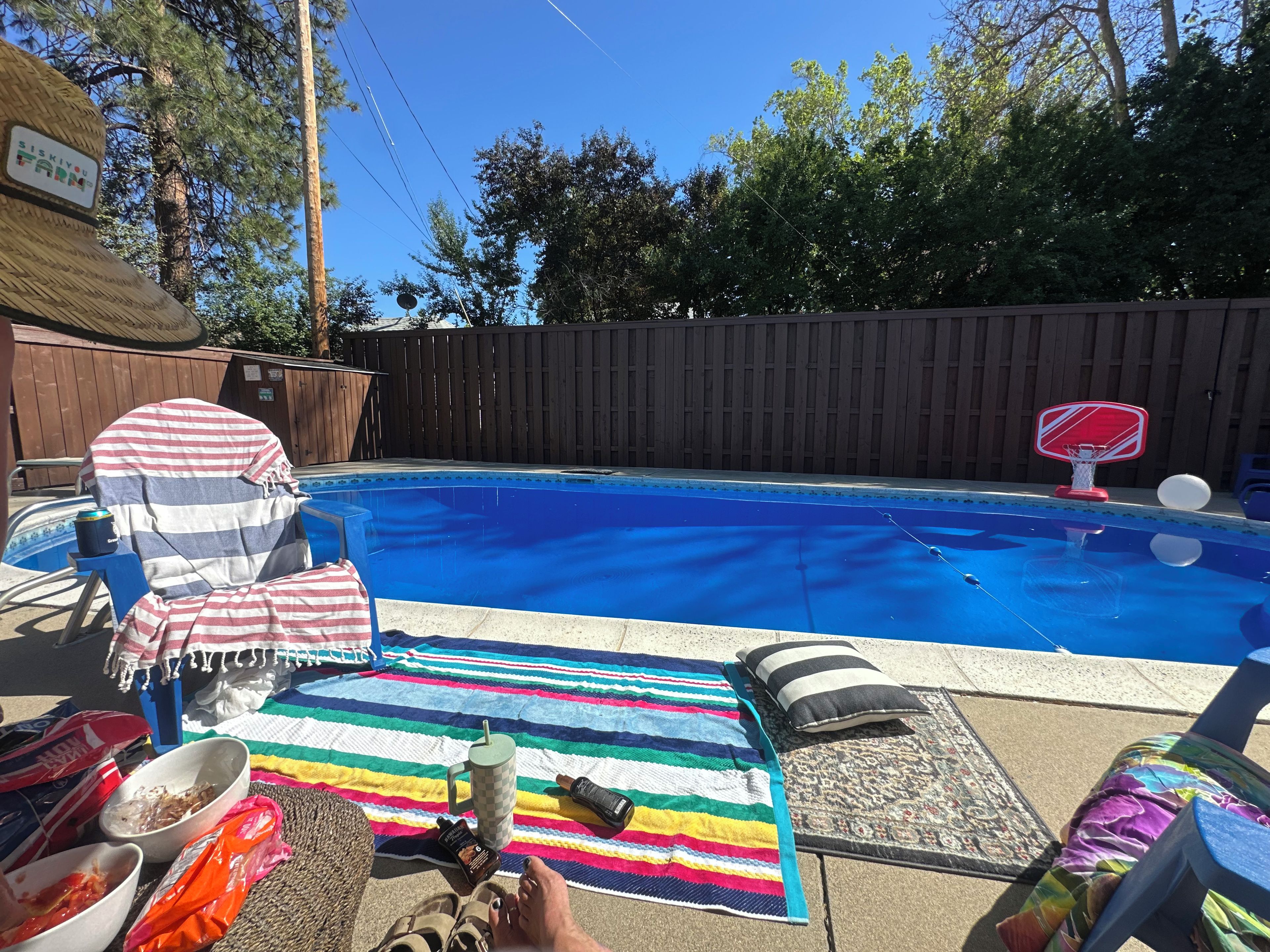 A backyard with a swimming pool, a wooden fence, and a lounge area with a striped blanket and a basketball hoop.