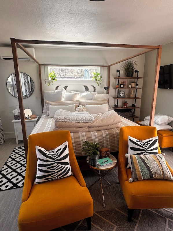 A modern bedroom with a four-poster bed, two armchairs, a bedside table with a lamp, a round mirror, and a bookshelf.