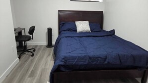 A bedroom with a bed, a desk, a chair, and a standing fan. - The Cozy Corner HOme (Winnipeg)