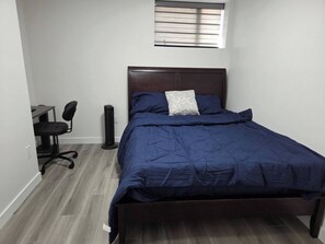 Een slaapkamer met een bed, een bureau, een stoel en een staande ventilator.