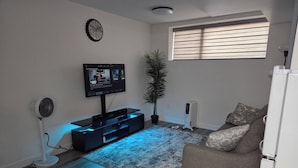 A living room with a flat-screen TV, a standing fan, a potted plant, a radiator, and a window with blinds. - The Cozy Corner HOme (Winnipeg)