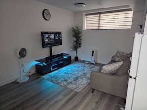 Een woonkamer met een flatscreen televisie, een ventilator, een potplant, een radiator en een raam met jaloezieën.