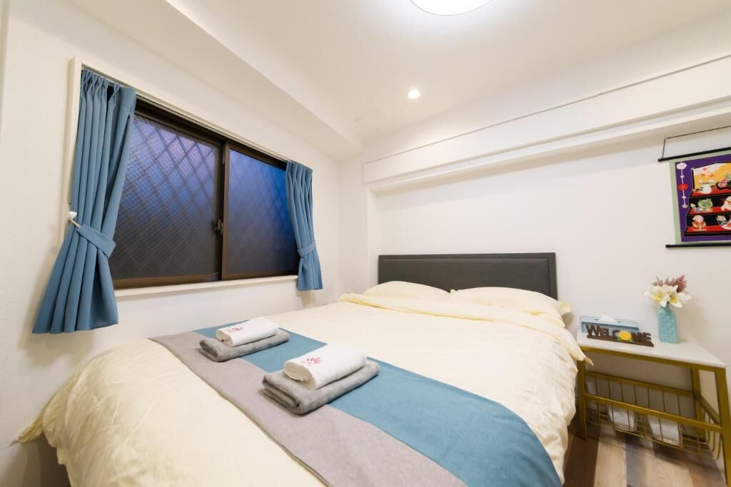 A neatly made bed with a blue and white bedspread, a grey blanket, and two folded towels. There is a window with blue curtains, a small bedside table with a vase of flowers, and a wall-mounted picture.