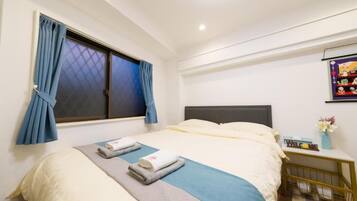 A neatly made bed with a blue and white bedspread, a grey blanket, and two folded towels. There is a window with blue curtains, a small bedside table with a vase of flowers, and a wall-mounted picture.