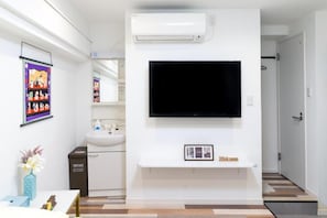 A modern hotel room with a mounted TV, a white console table with a welcome sign, a bathroom with a sink and mirror, and a container for toiletries.