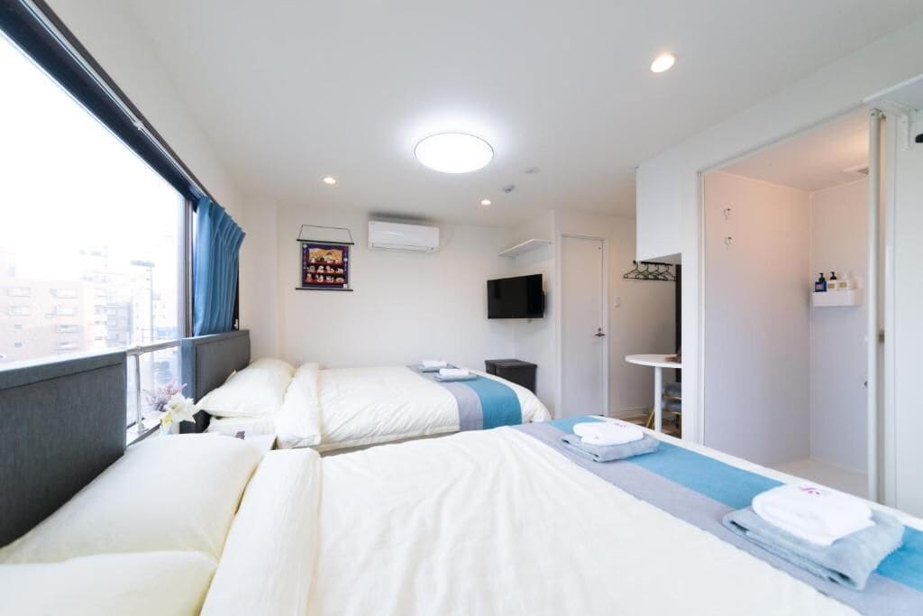 Habitación de hotel con dos camas, televisión, ventana con cortinas, puerta que da al baño y una mesita con artículos de aseo.