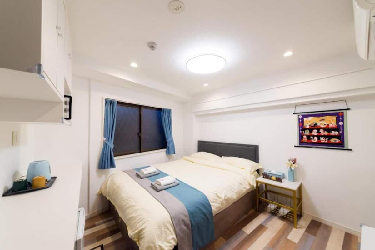 Un dormitorio con cama, mesita de noche, ventana con cortinas e imagen colgada en la pared.