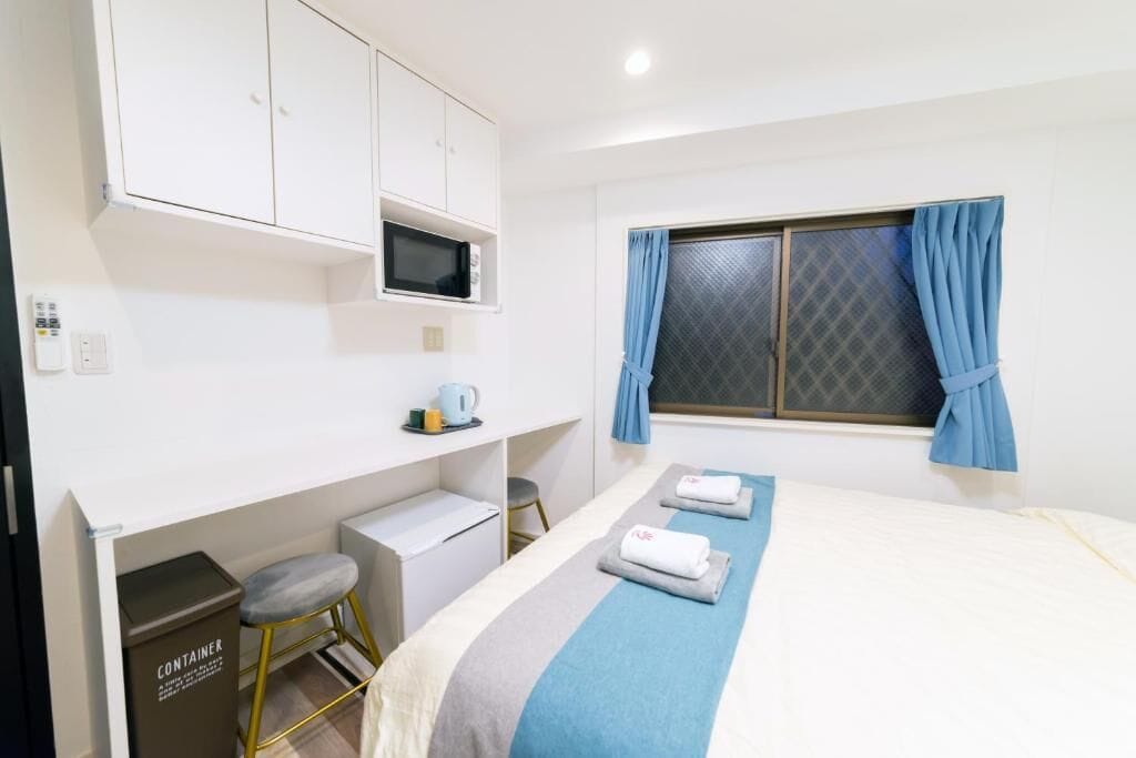 A hotel room with a bed, a desk with a chair, a microwave, a trash bin, and a window with curtains.