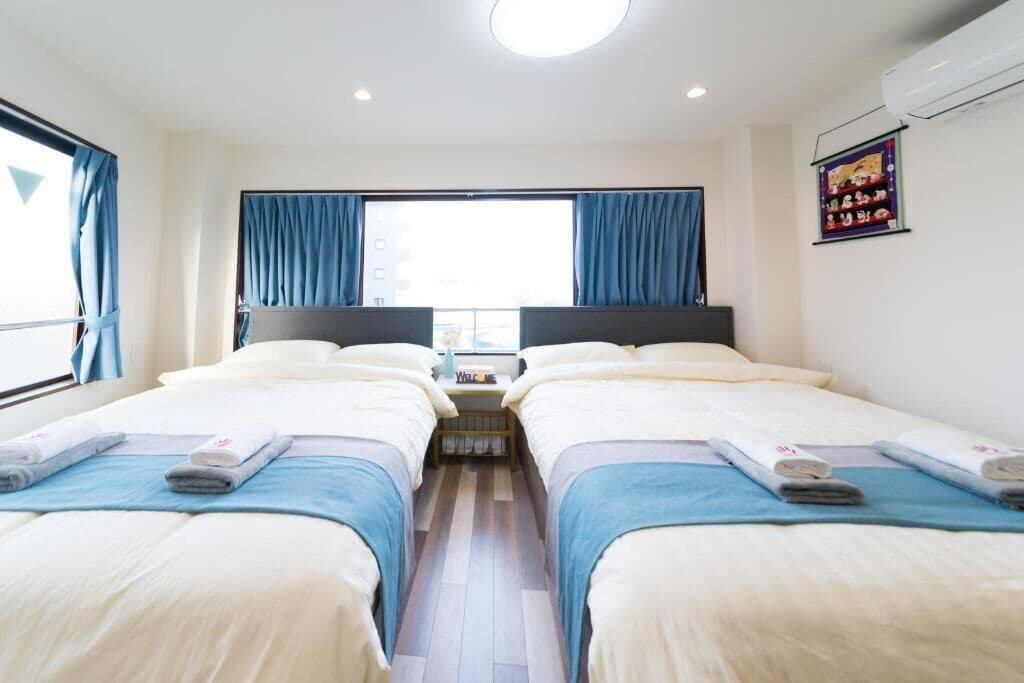 A hotel room with two beds, a window with curtains, a small table with a lamp, and an air conditioning unit on the wall.