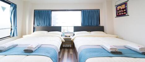 A hotel room with two beds, a window with curtains, a small table with a lamp, and an air conditioning unit on the wall.