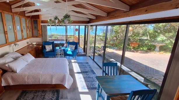 Une chambre à coucher avec un lit, une petite table, deux chaises et une vue sur la mer grâce à de grandes fenêtres.