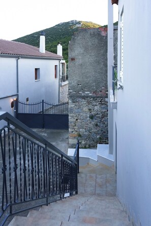 A narrow stairway leading to a stone wall, with a metal railing on one side and a white building on the other.