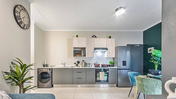 A modern kitchen with a washing machine, a dining area with a blue chair, and a clock on the wall. - Zalari Luxe 93 on New, 1 bed, Central & Premium Comfort (Midrand)