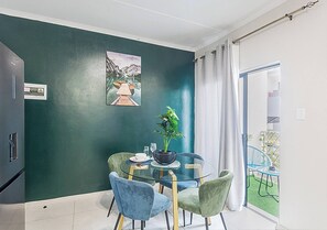 Uma sala de jantar com mesa de vidro, cadeiras, um vaso de planta e um quadro na parede.