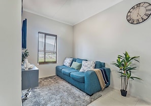 Ein Wohnzimmer mit einem blauen Sofa, einer Uhr, einem Fenster und einer Topfpflanze.