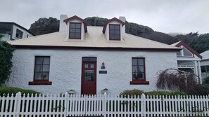 Una casa de poble blanca amb una porta vermella, dues finestres laterals inclinades i una tanca de palets blanca.
