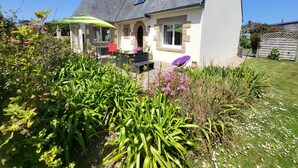 Ein Haus mit geneigtem Dach, einer Terrasse mit Gartenmöbeln und einem Garten mit verschiedenen Pflanzen und Blumen.