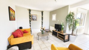 Ein Wohnzimmer mit gelber Couch, einem kleinen runden Tisch, einer TV-Konsole und einem Topf mit Pflanze.
