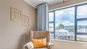 Une pièce avec une grande fenêtre, un fauteuil avec un coussin et deux œuvres d’art encadrées accrochées au mur.