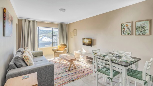 Ein modernes Wohnzimmer mit einer Couch, einem Esstisch für vier Personen, einem an der Wand befestigten Fernseher und einem großen Fenster mit Vorhängen.