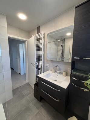 Une salle de bain moderne avec un lavabo blanc, des meubles de rangement aux couleurs foncées et une cabine de douche en verre.