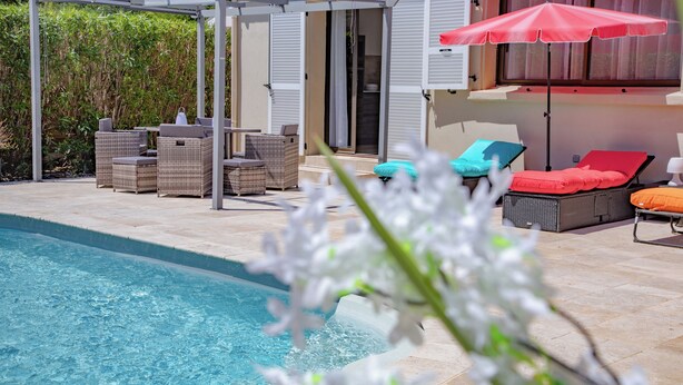 Un área junto a la piscina con mobiliario de mimbre, una pérgola y un paraguas rojo.