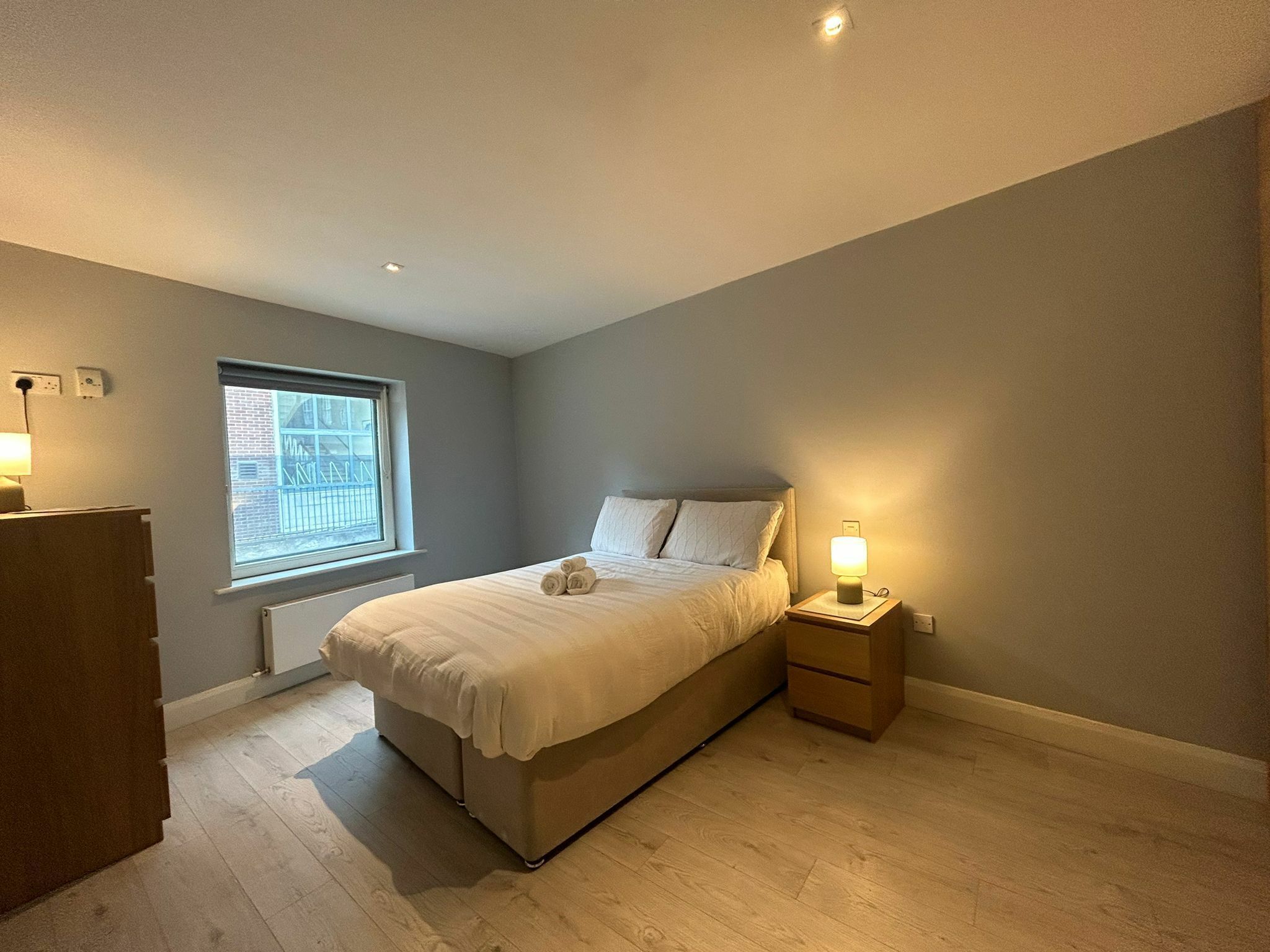 Un dormitorio con cama, mesita de noche, lámpara y ventana.