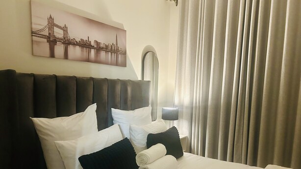 A hotel room with a bed, pillows, a headboard, a nightstand with a lamp, and a window with curtains. - Cozy Apartment in the heart of Joburg. (Midrand)
