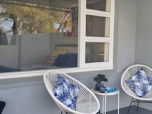 Quarto com uma grande janela, duas cadeiras de vime brancas com almofadas estampadas em azul, uma mesinha com um vaso de planta e uma cama com almofadas listradas.