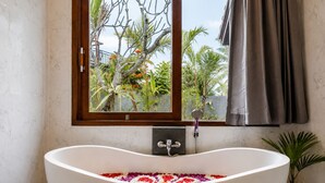 A bathroom with a freestanding bathtub, a window with a view of tropical plants, and a showerhead. - Togog Sebatu Villa (Gianyar)