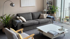 Una sala de estar con un sofá gris, una silla de madera, una mesa de centro blanca, una lámpara de pie, una estantería y un ventanal grande.