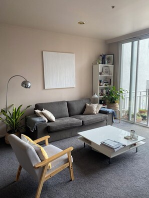 A living room with a grey sofa, a wooden chair, a white coffee table, a floor lamp, a bookshelf, and a large window. - Apartment in Melb's Heart (Southbank)