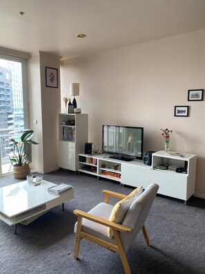 A modern living room with a flat-screen TV, white entertainment unit, a potted plant, and a comfortable chair. - Apartment in Melb's Heart (Southbank)