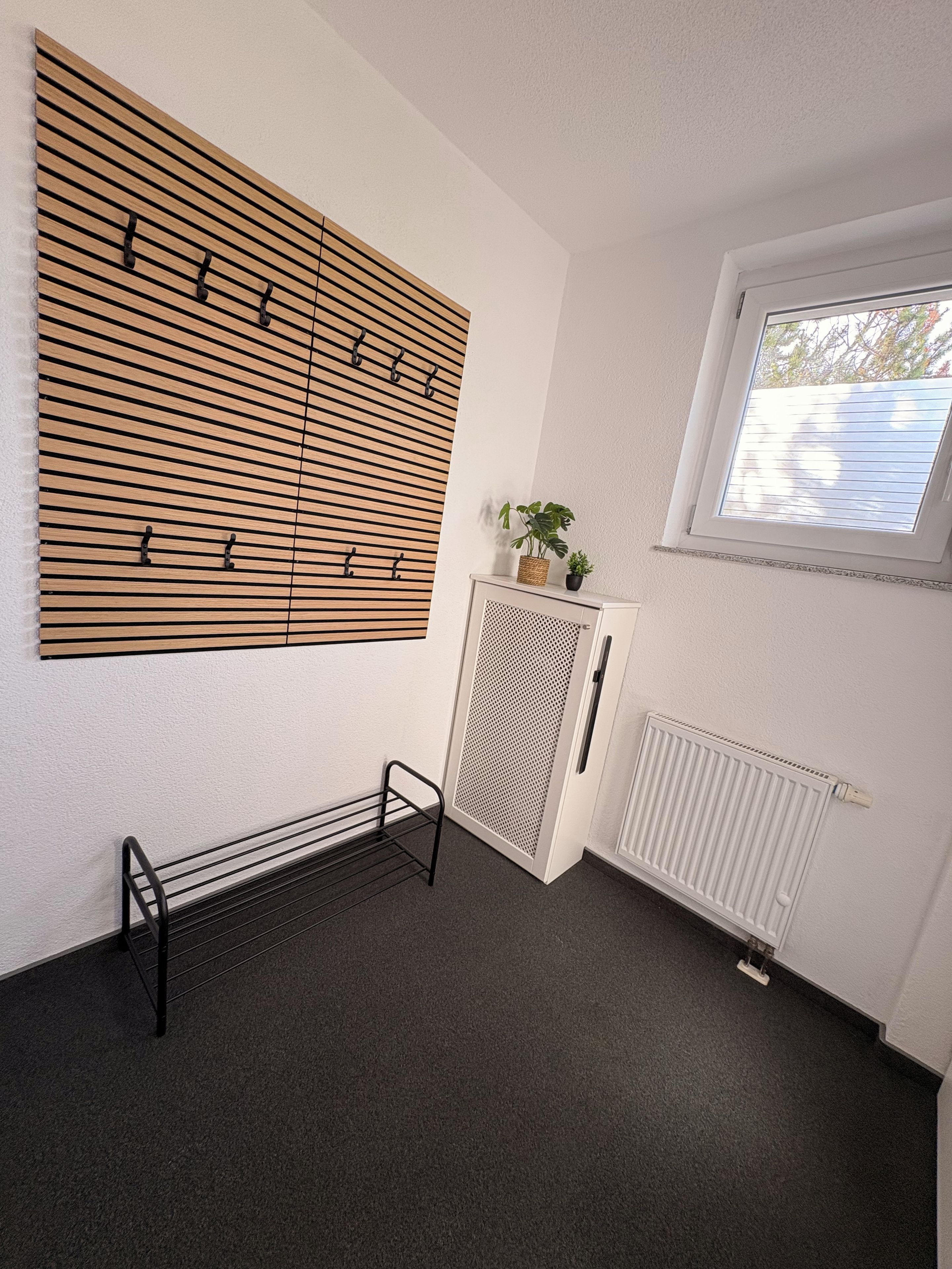 Ein Zimmer mit einer durchbrochenen Holzwand, einem Heizkörper, einem Fenster mit Jalousien, einem kleinen Schrank mit einer Pflanze und einem Metallregal.