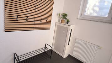 Ein Zimmer mit einer durchbrochenen Holzwand, einem Heizkörper, einem Fenster mit Jalousien, einem kleinen Schrank mit einer Pflanze und einem Metallregal.