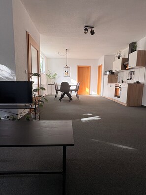 A modern kitchen with white cabinets, a built-in oven, and a microwave. There is a dining area with a table and chairs, and a TV on a stand.