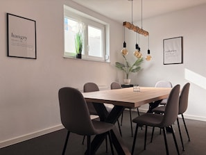 A modern dining area with a wooden table, six chairs, pendant lighting, and framed educational posters on the walls.
