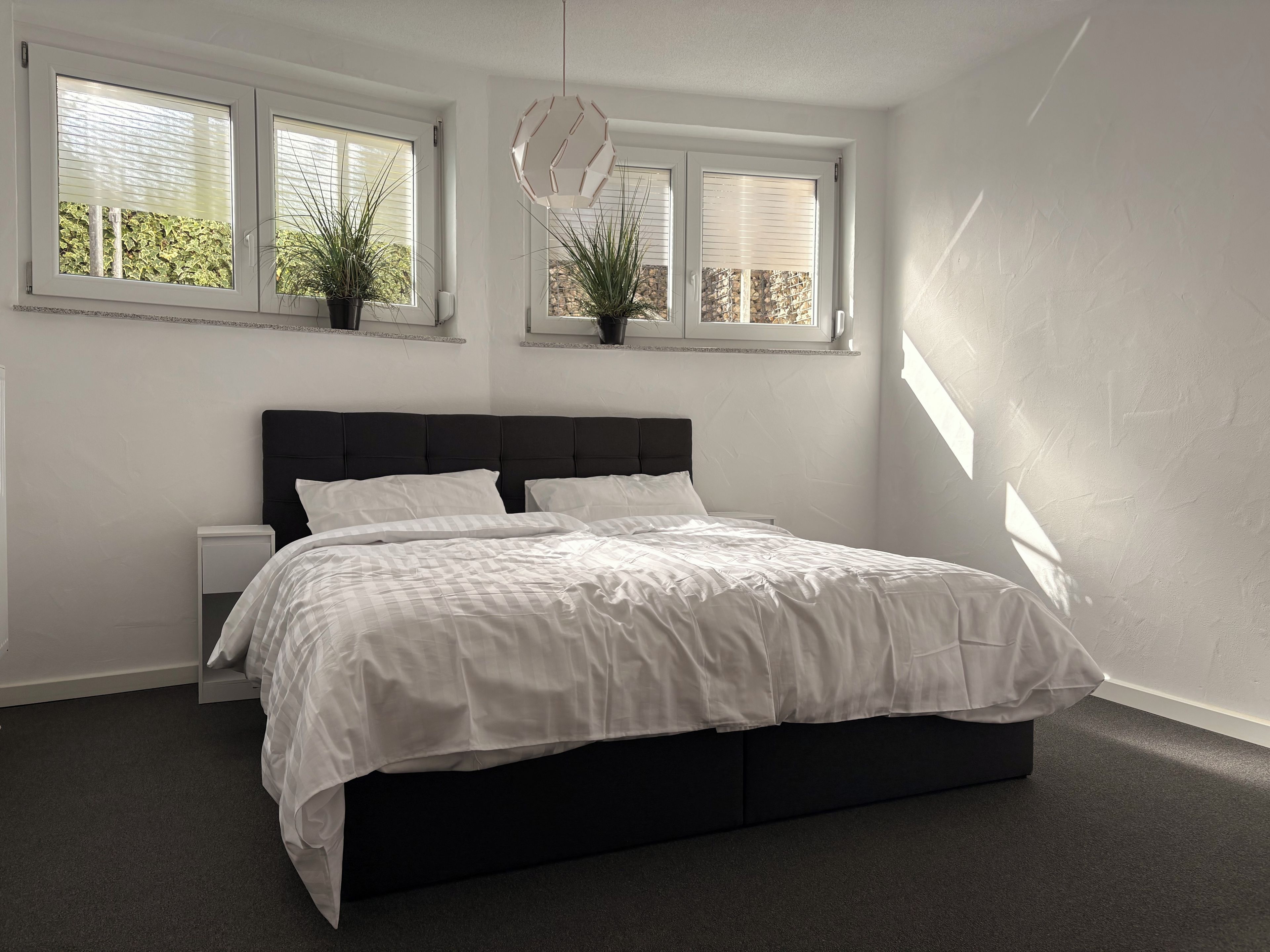 A bedroom with a large bed, two bedside tables, and three windows with plants.