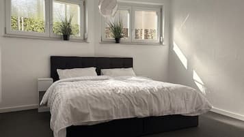 A bedroom with a large bed, two bedside tables, and three windows with plants.