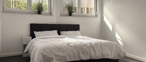 A bedroom with a large bed, two bedside tables, and three windows with plants.