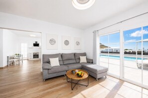 Ein modernes Wohnzimmer mit einer grauen Eckcouch, einem runden Couchtisch mit Früchten und einem großen Fenster mit Blick auf einen Pool.