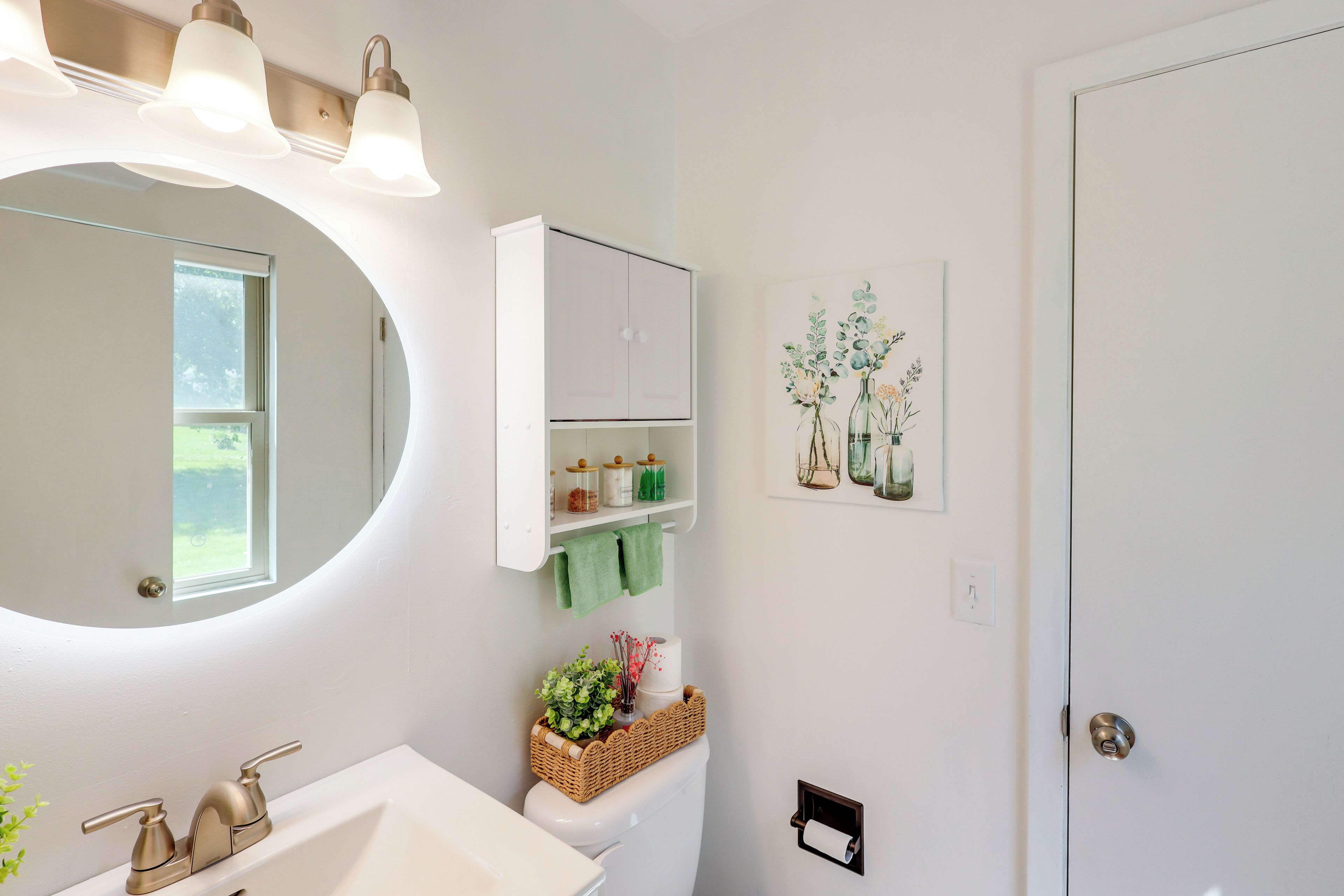 A bathroom with a white sink, a mirror with lights, a toilet with a basket of flowers, and a door.