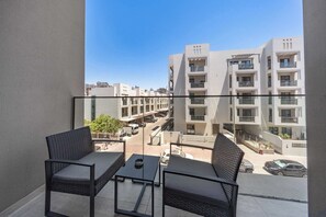 Ein Balkon mit zwei Korbstühlen und einem kleinen Tisch, mit Blick auf eine Straße mit Gebäuden und parkenden Autos.