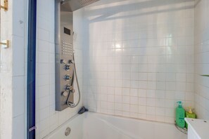 Een badkamer met witte betegelde muren, een bad, een douchekop en een plank met toiletartikelen.